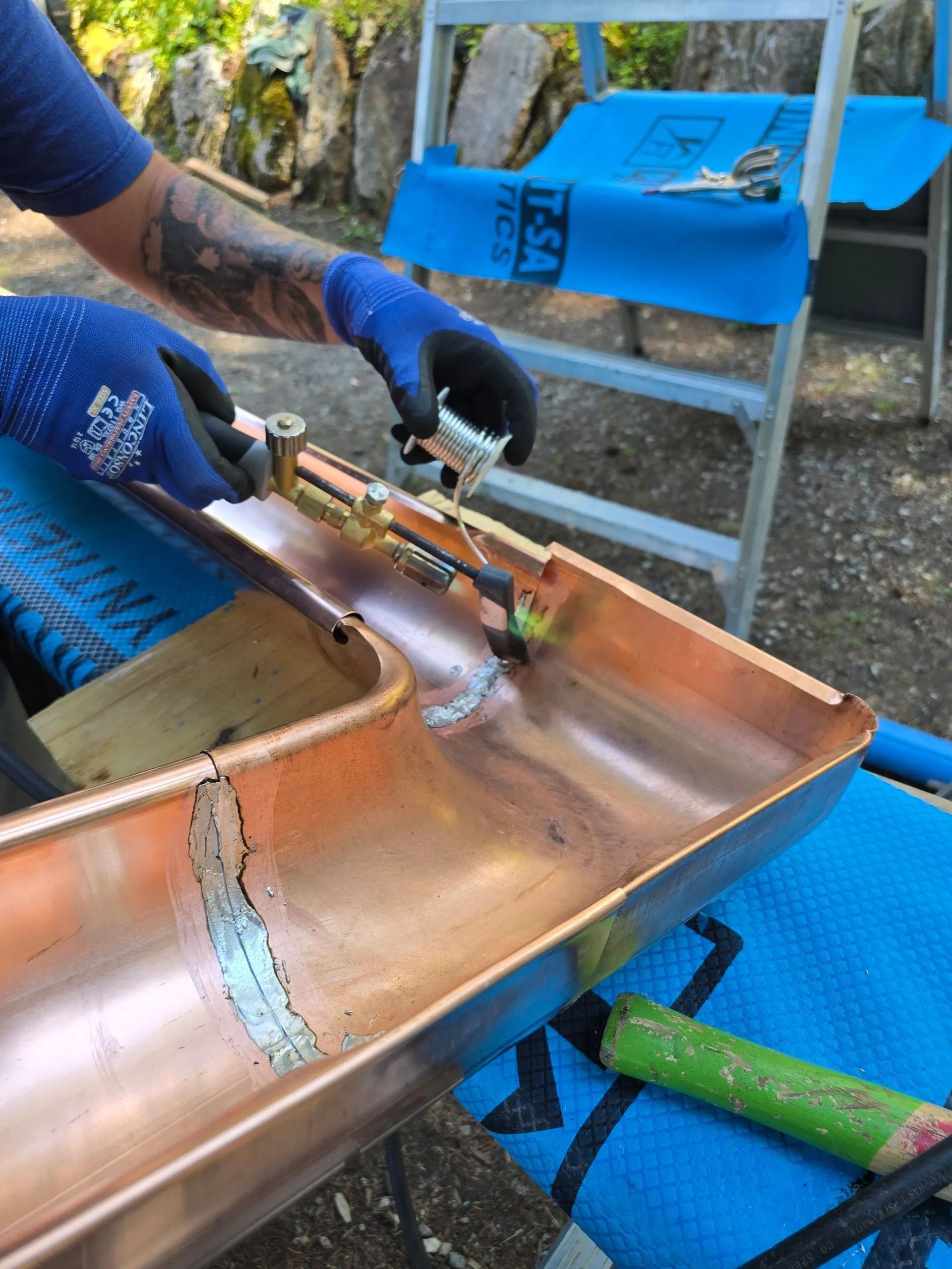 Worker welding a copper gutter on a ladder, with blue gloves and tools nearby.