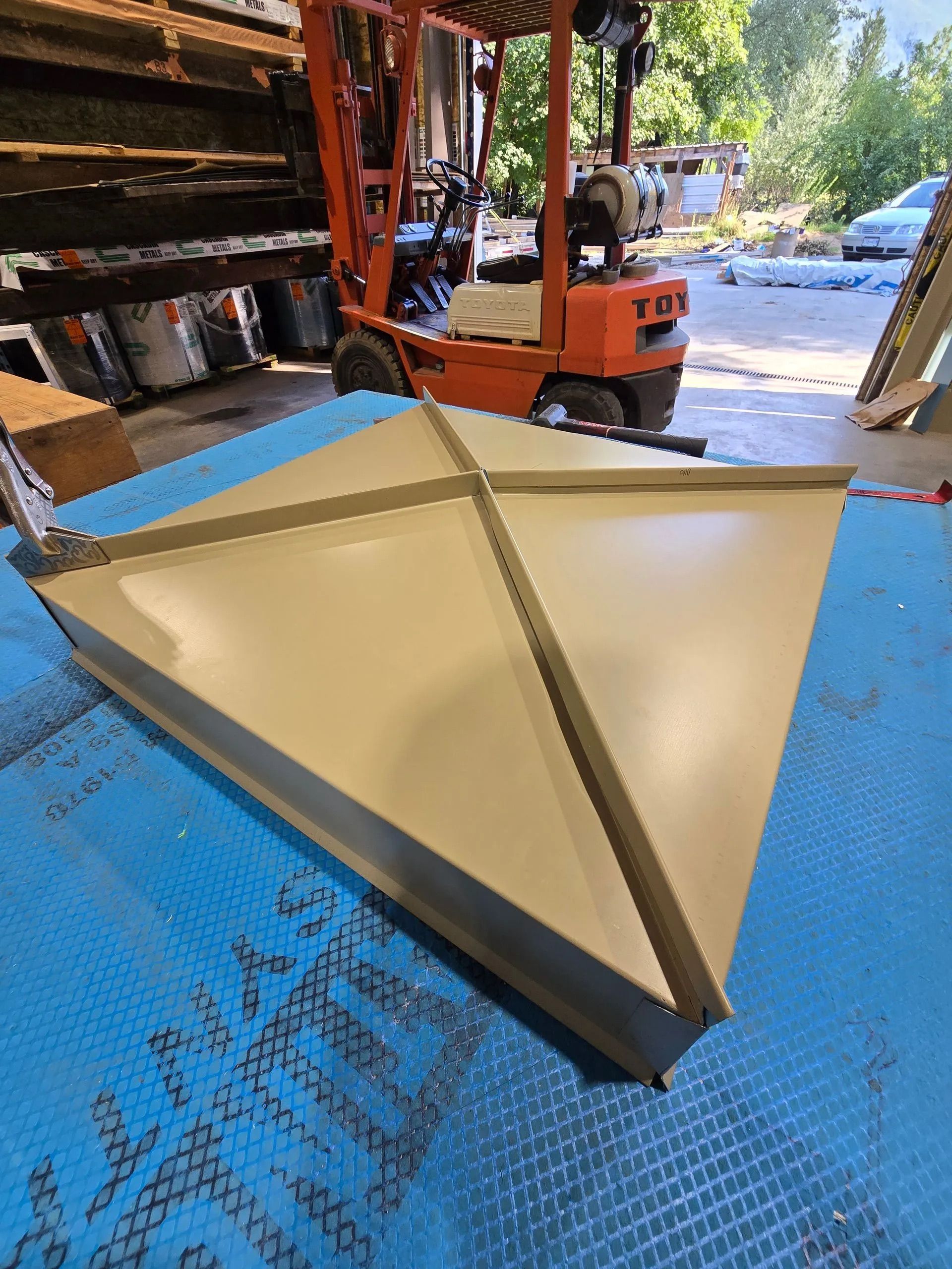 Beige triangular metal roof panel on a blue floor in a workshop, with an orange forklift nearby