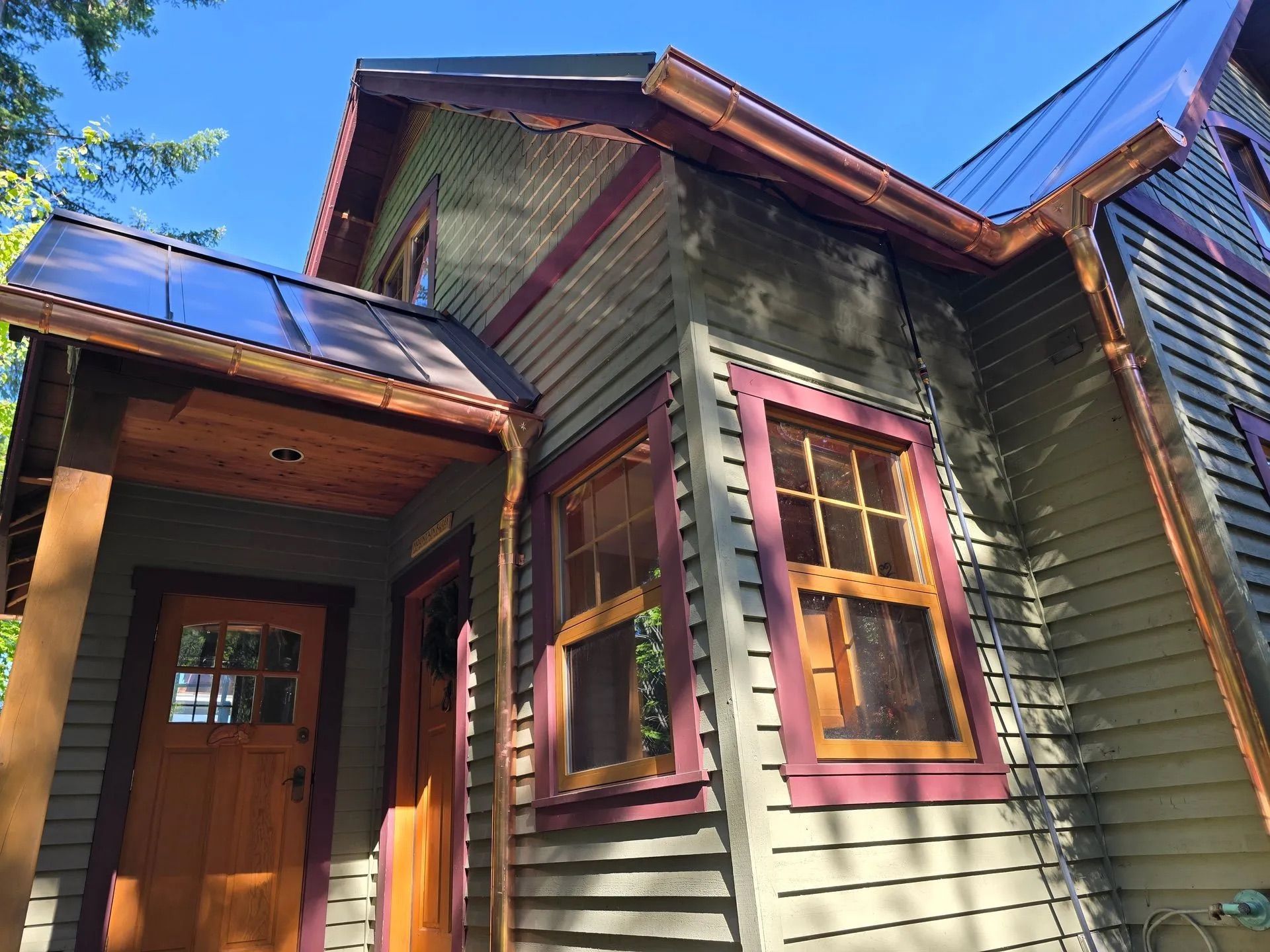 Exterior of a rustic wooden house with green siding, red trim, and a blue metal roof under a clear sky