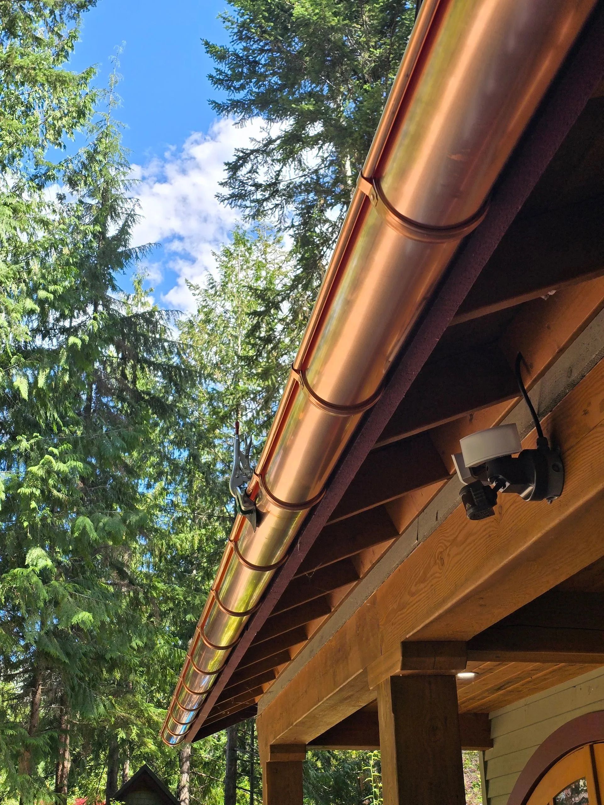 Wooden cabin eaves with copper gutters under tall evergreen trees and a blue sky