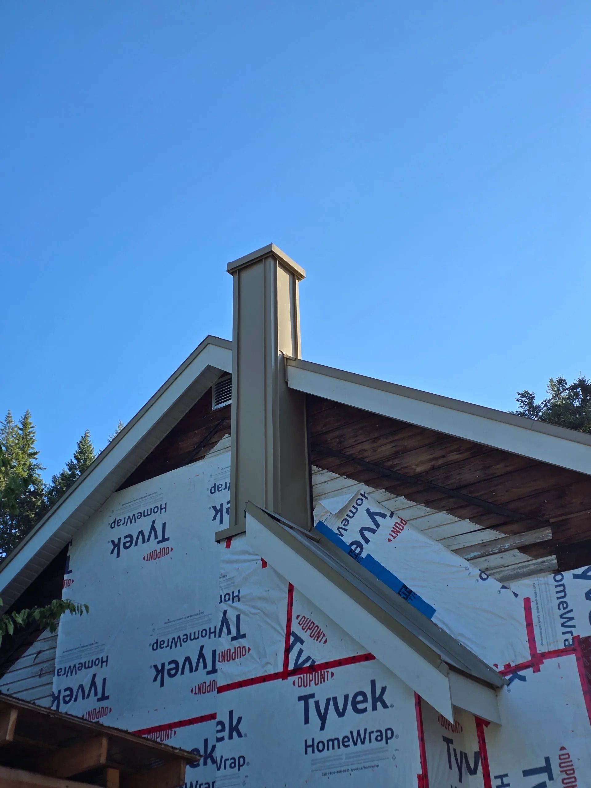 House exterior under construction with Tyvek wrap and a roof chimney against a blue sky