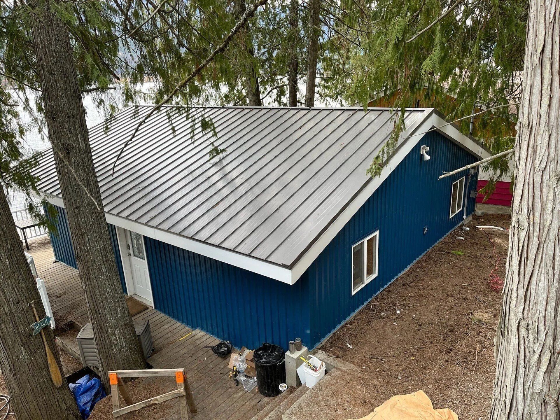 Blue house with a gray metal roof among trees, seen from above, with outdoor items near the wall