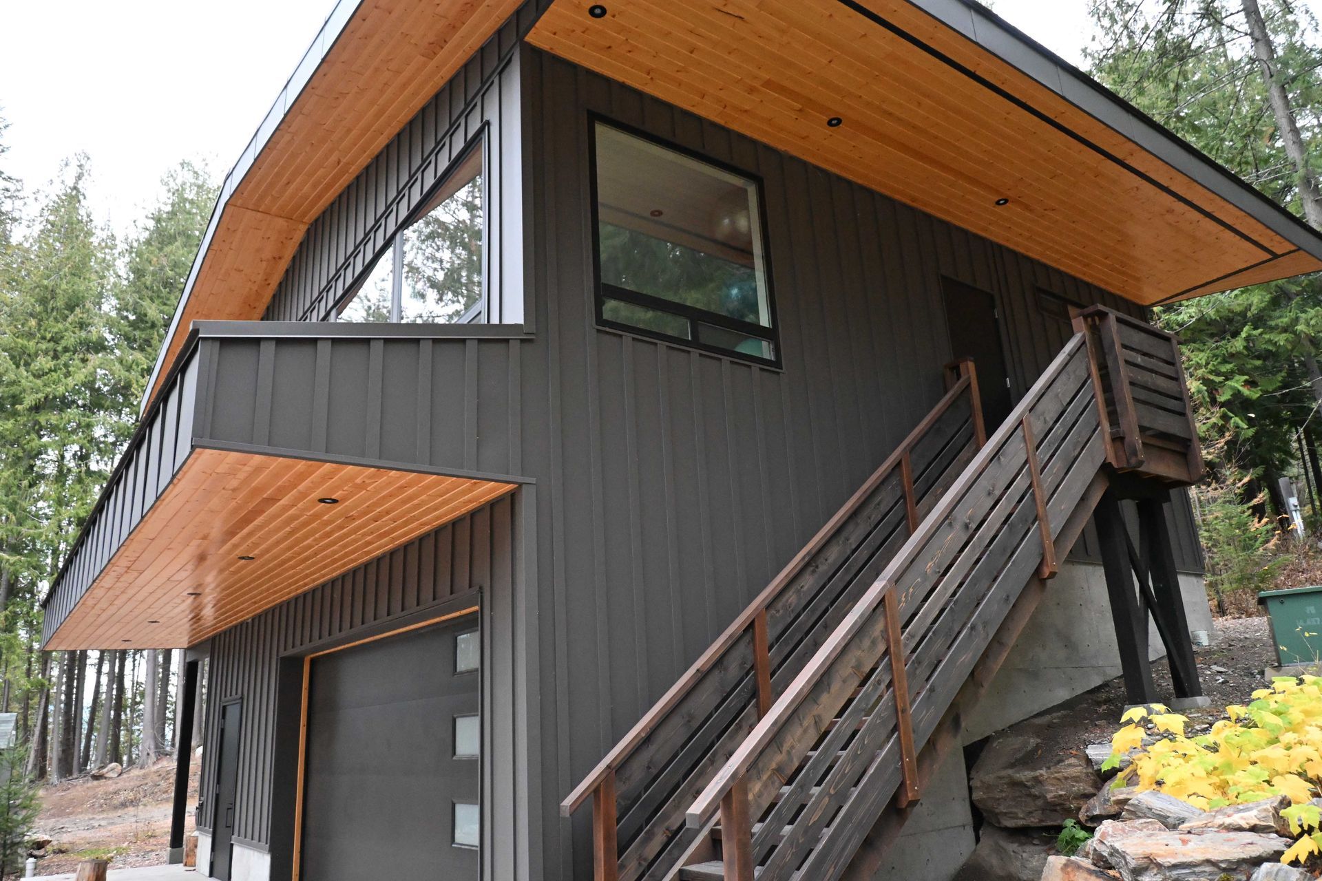 Modern dark-gray house with wood soffits and an exterior staircase in a forest setting