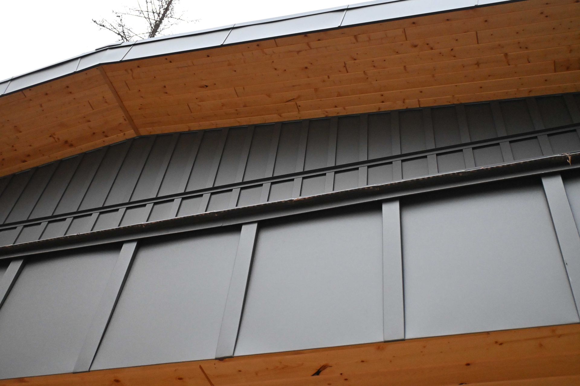 Close-up of a modern building facade with gray panels and a wooden roof overhang.