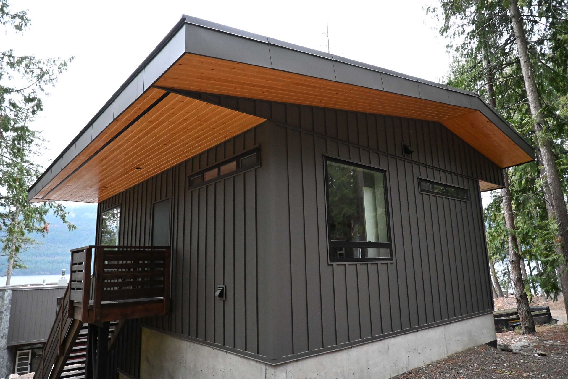 Modern dark-gray cabin with a wood soffit, covered porch, and large window in a wooded area