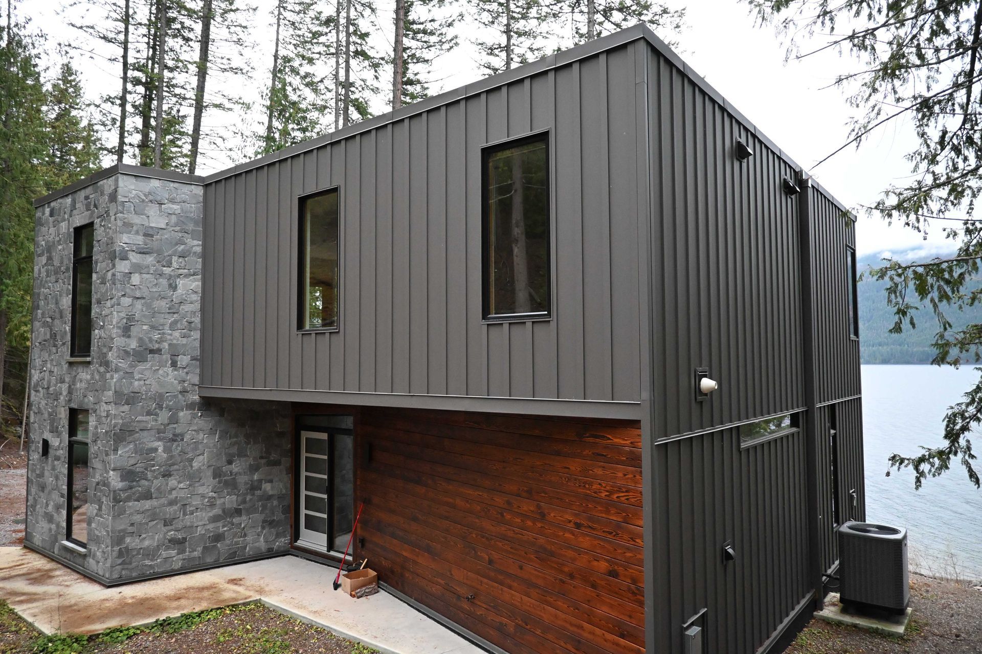 Modern two-story house with gray metal siding, stone wall, and wood accent in a forested setting