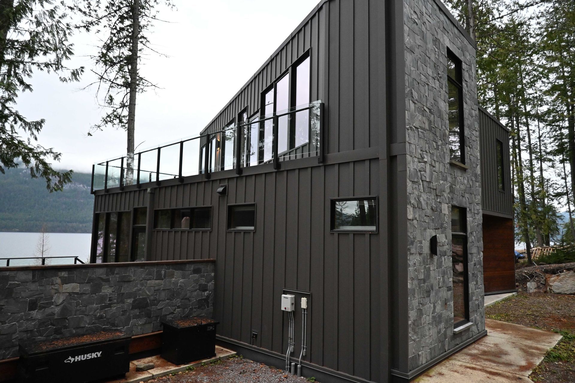 Dark gray modern house with stone accents and a rooftop deck beside a wooded lakeside.