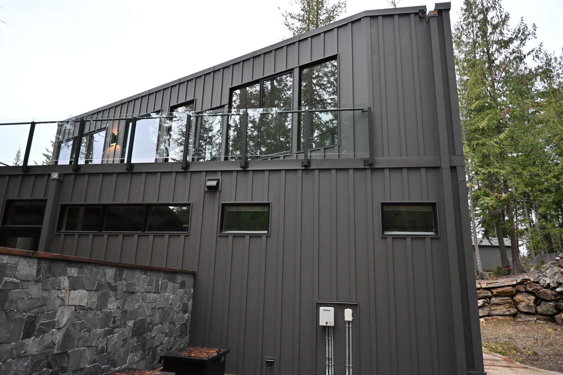 Modern dark gray metal house with large windows, stone retaining wall, and trees in the background