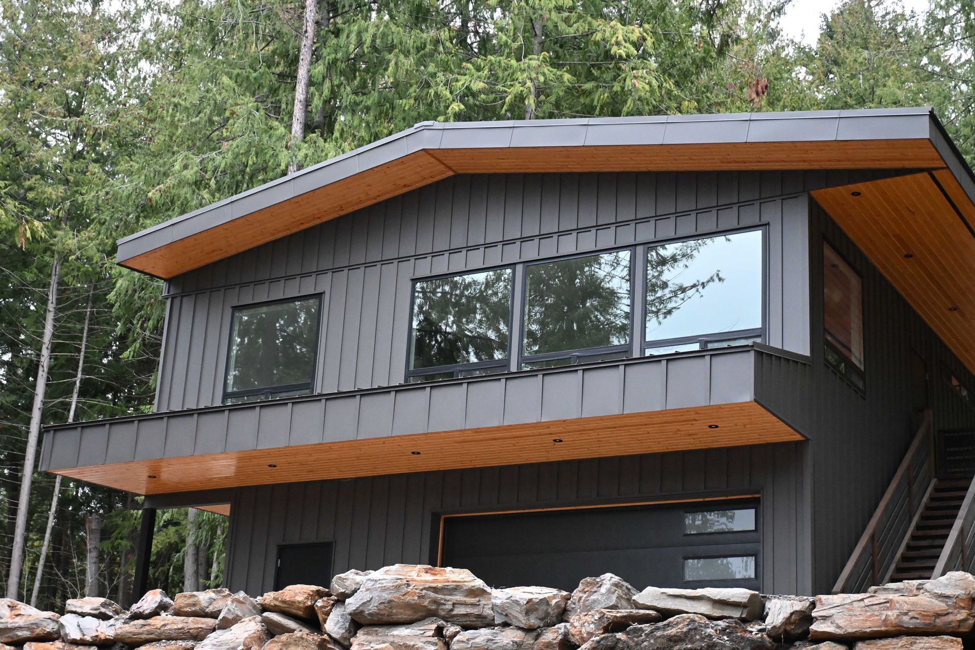 Modern dark-gray house with wood trim and large windows on a rocky hillside surrounded by trees