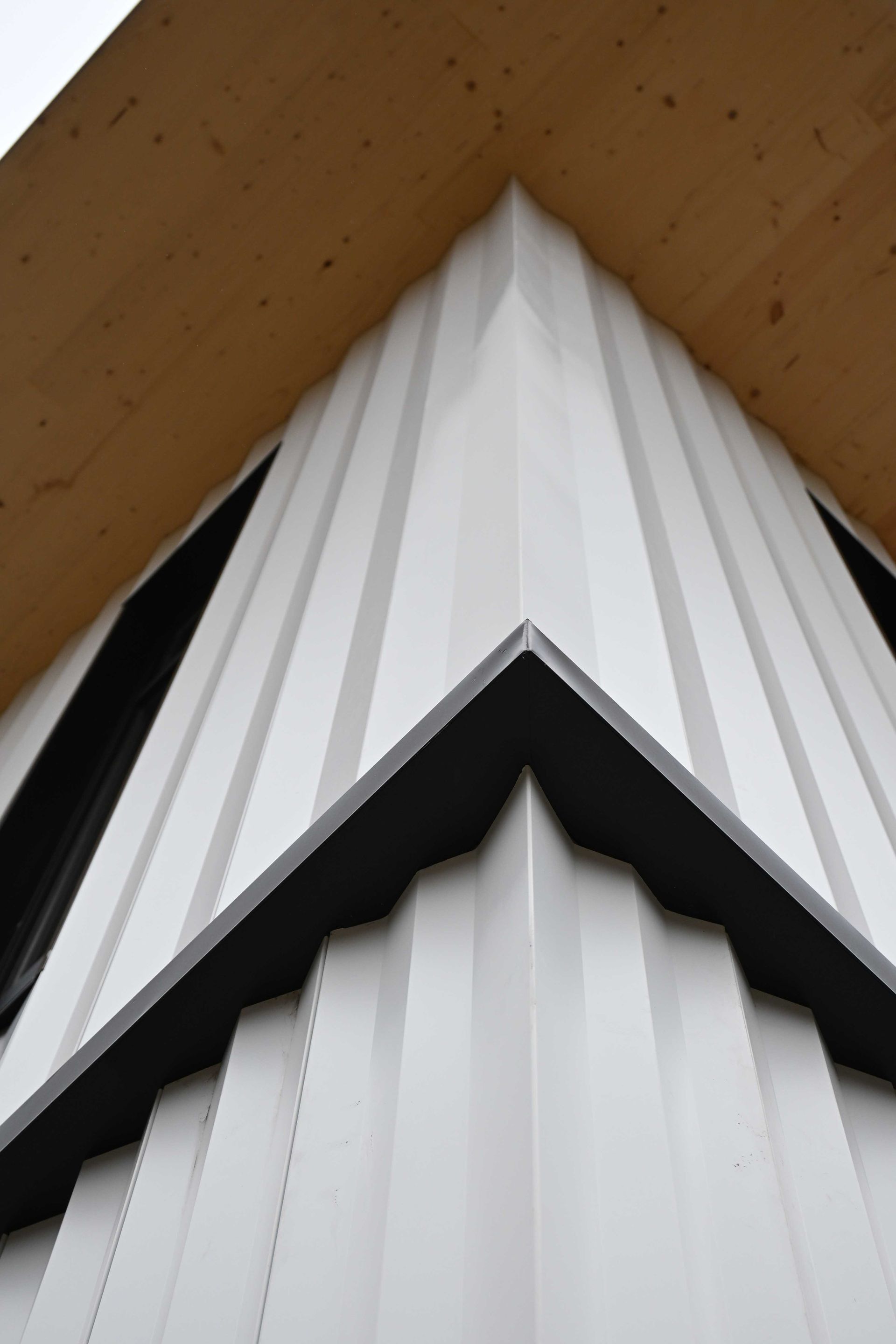 White pleated architectural panels meeting at a sharp black triangular seam under a wood ceiling