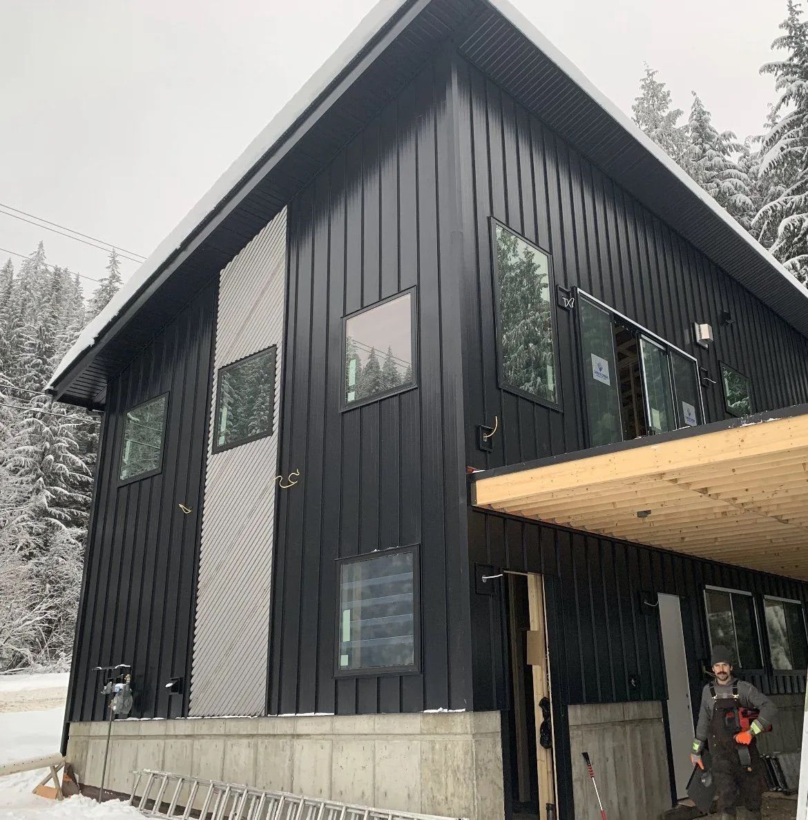 Black modern cabin with large windows, concrete foundation, and a covered deck in a snowy forest setting