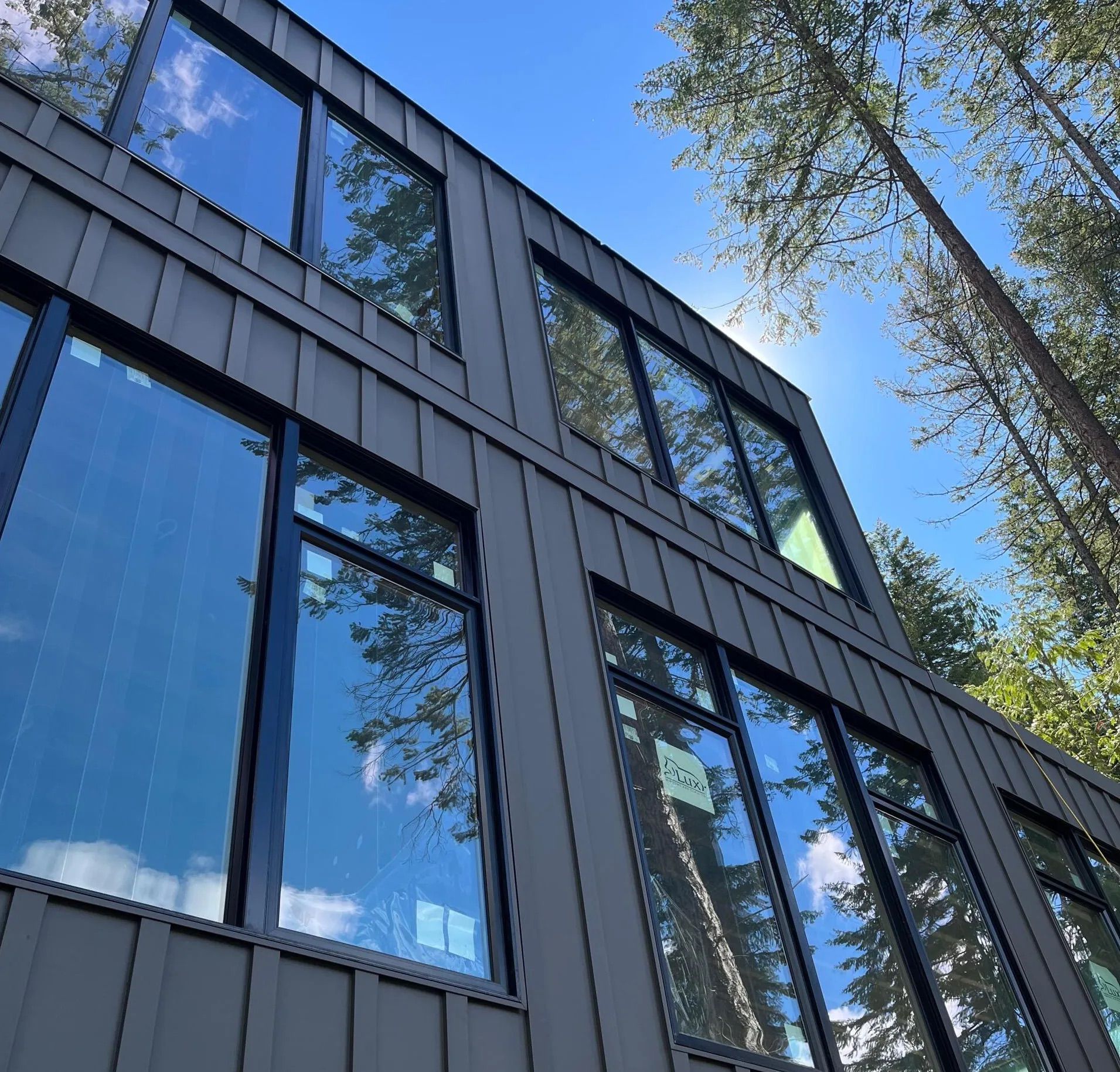 Modern dark-gray building with large reflective windows viewed from below against blue sky and trees