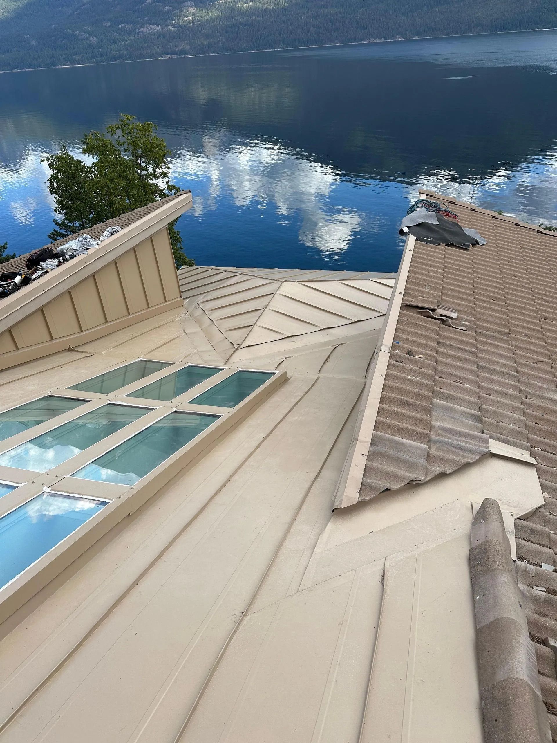 Rooftop view of a steep, tan shingled roof beside a blue lake with clouds reflected in the water.