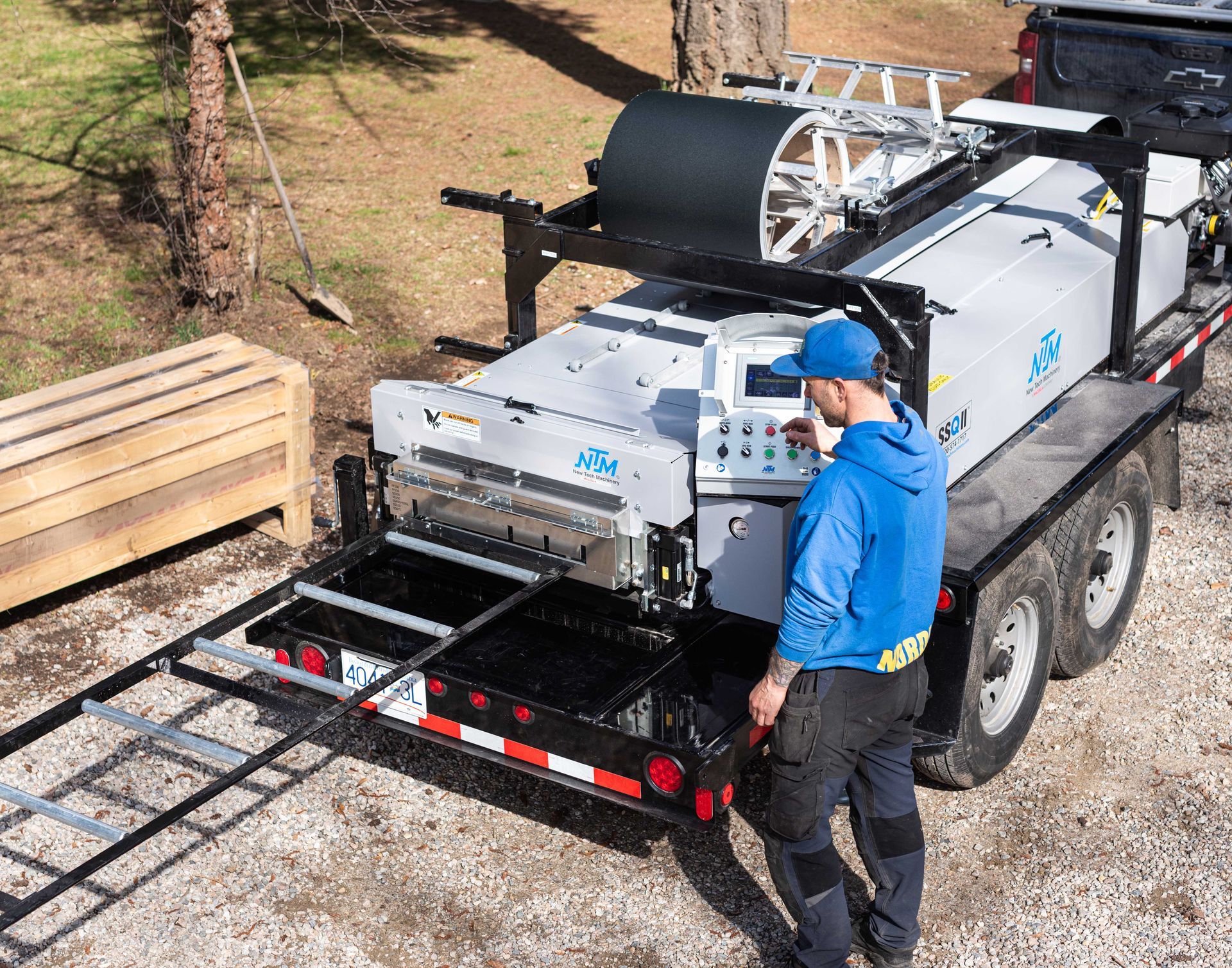 Worker in blue jacket inspecting a large industrial machine on a trailer in a gravel yard.