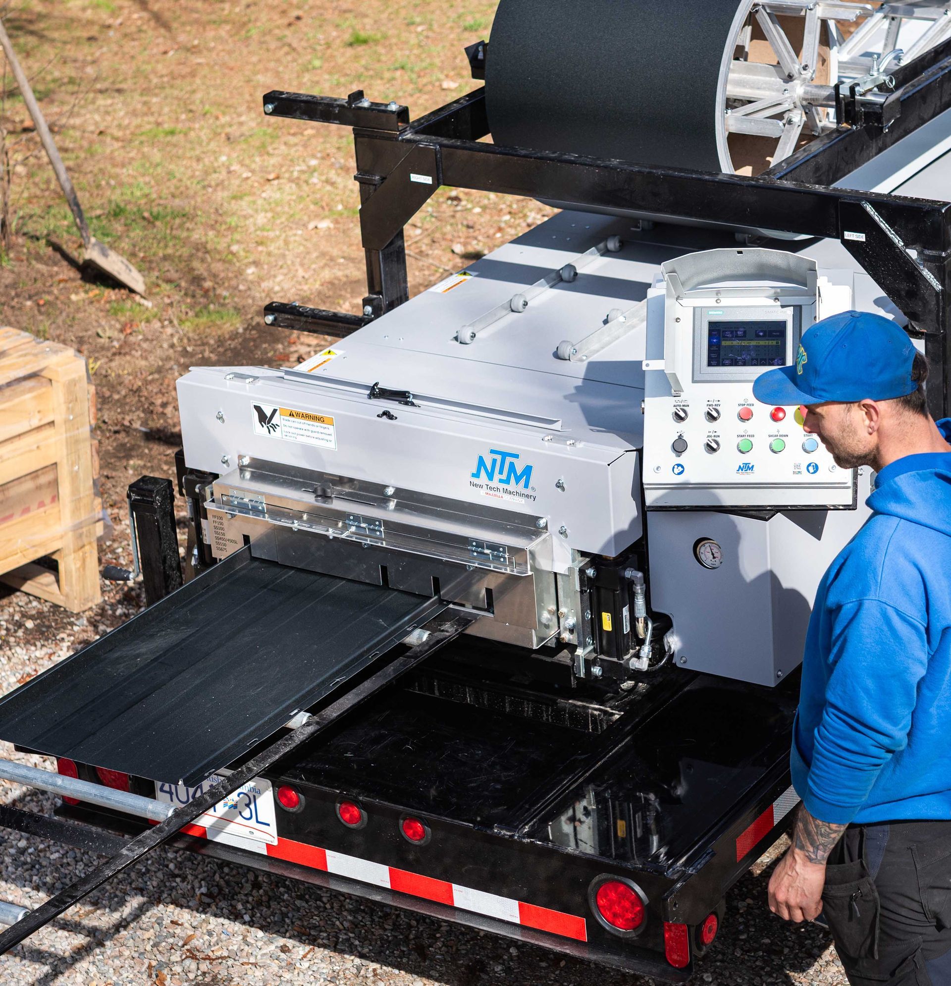 Large silver industrial machine on a trailer, with a worker in blue beside it outdoors.