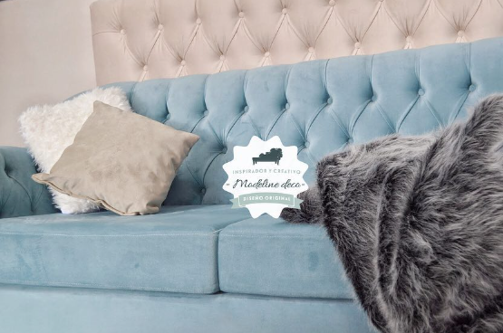 Sofá de terciopelo azul con respaldo capitoné, almohadones beige y una manta de piel sintética gris.