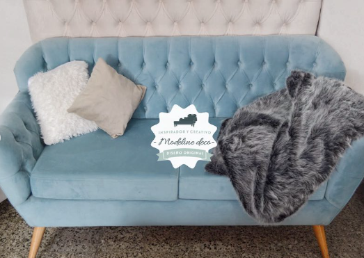 Sofá de terciopelo azul con respaldo capitoné, almohadones beige y una manta de piel sintética gris.