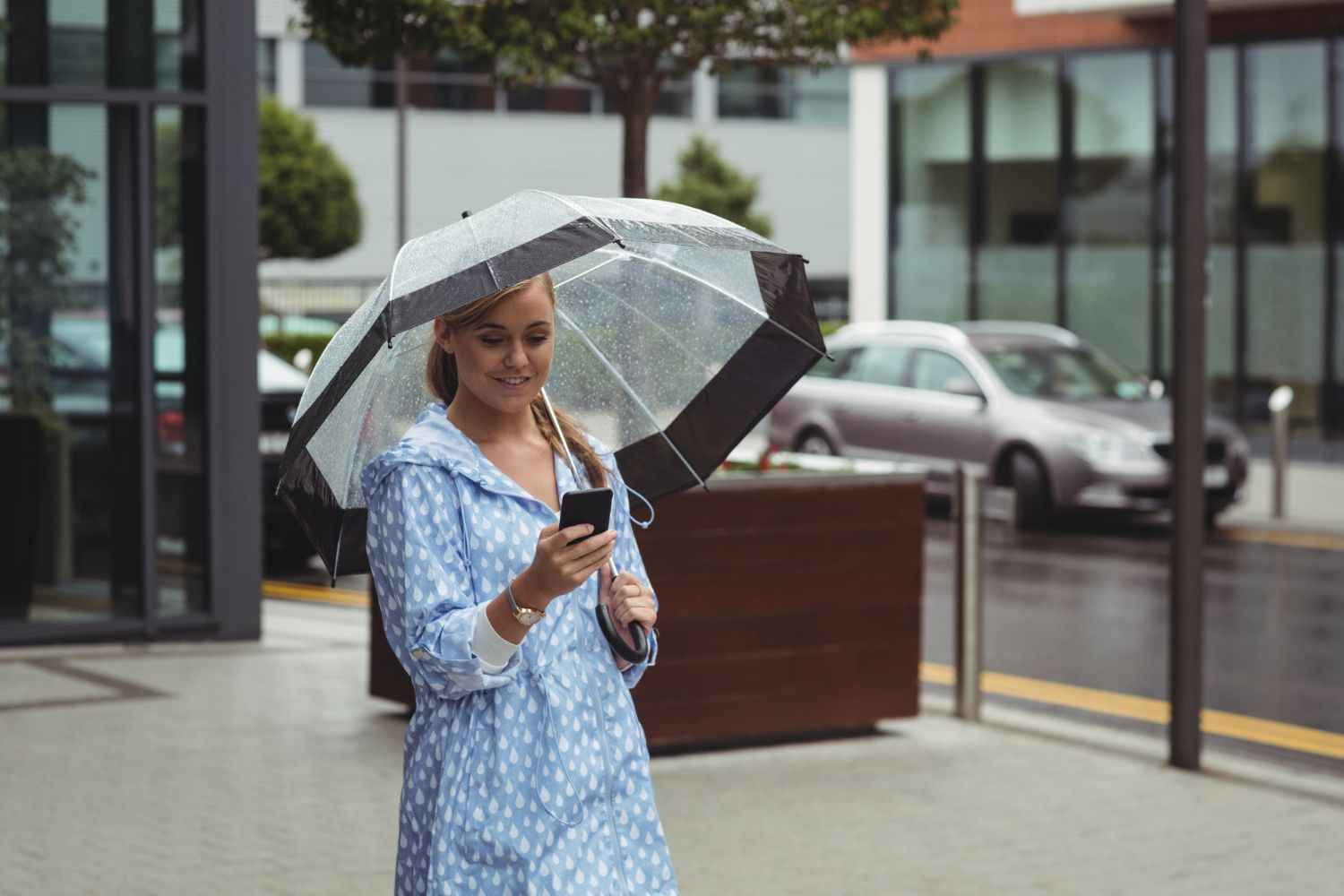 A person in a blue patterned coat walks outside holding an umbrella while looking at a smartphone.