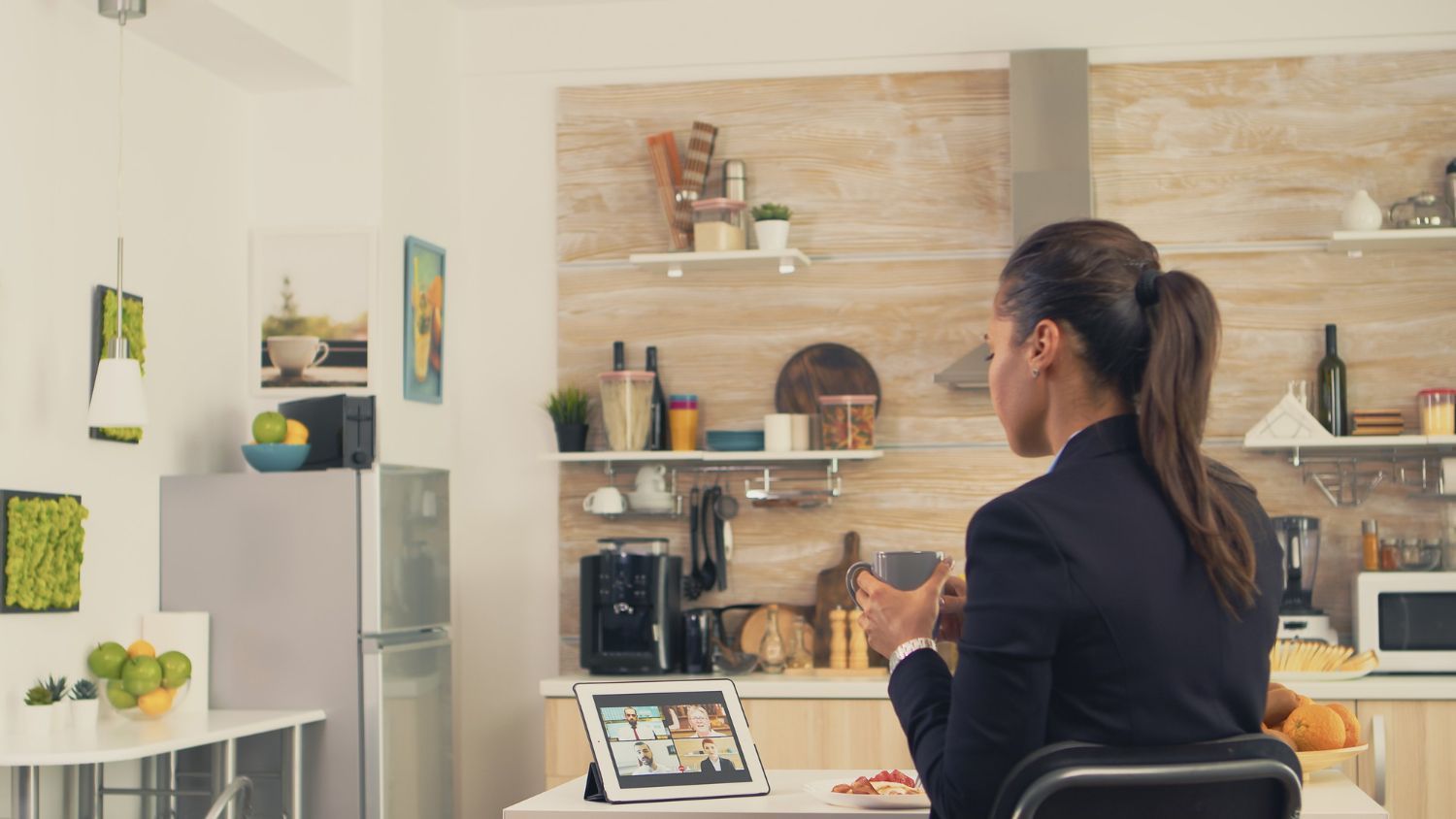 A person in a business blazer sits at a kitchen table, participating in a video conference on a tablet.