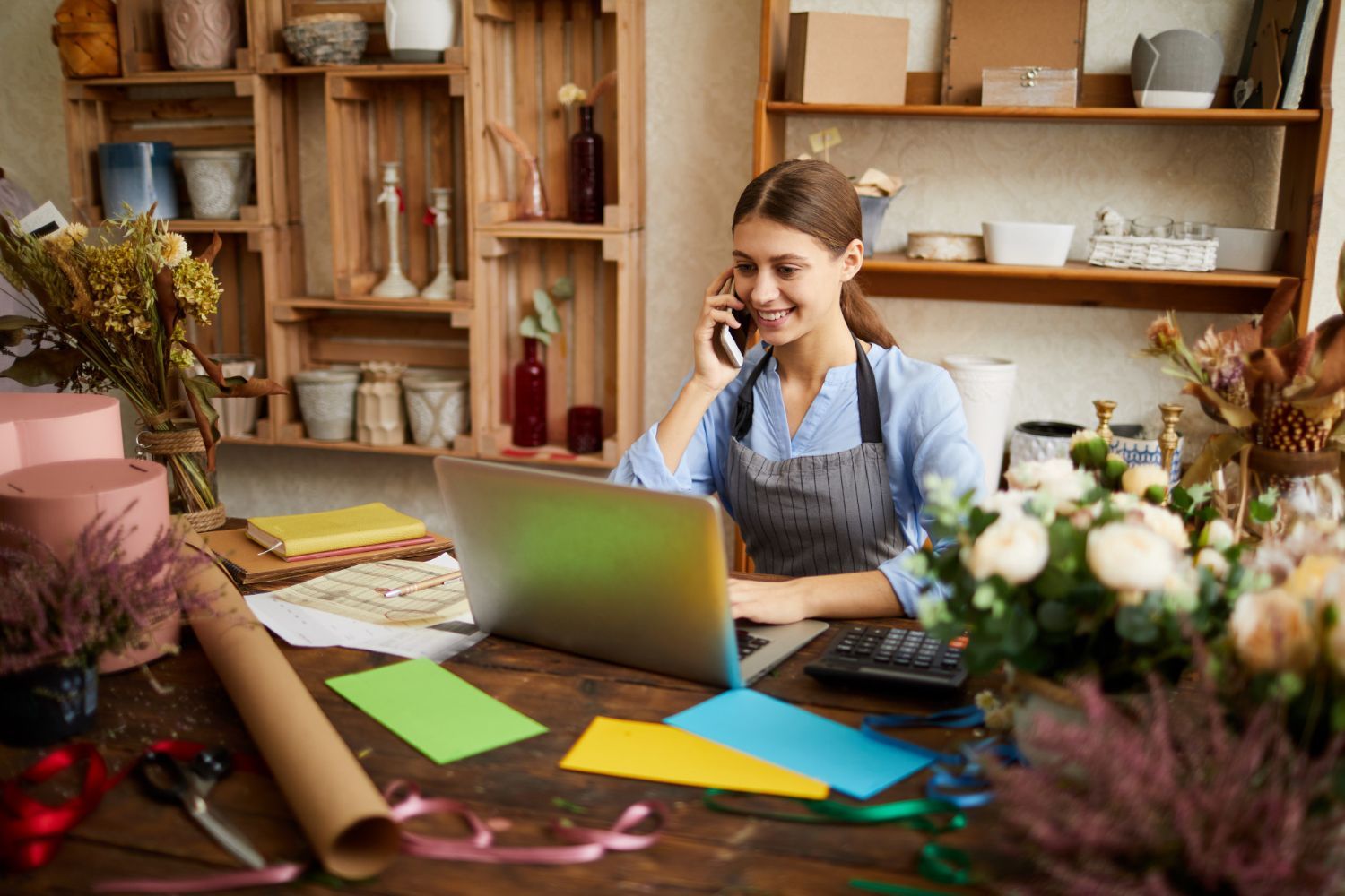 A person in an apron talks on the phone while using a laptop at a flower shop desk filled with ribbons and floral supplies.