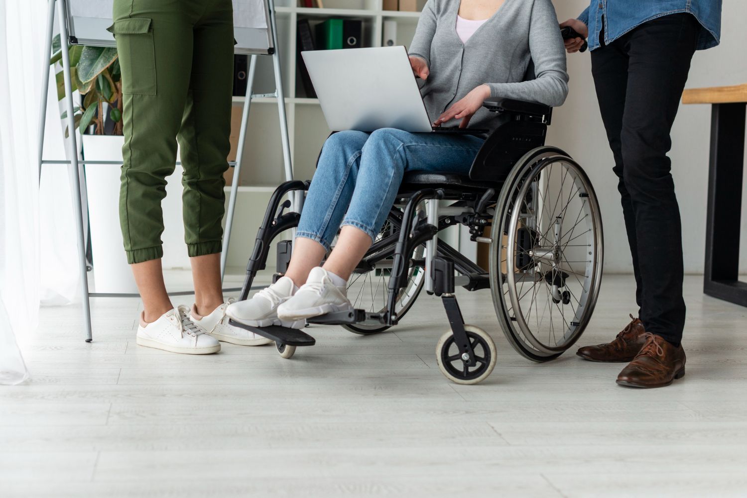 Three people stand in an office; one person in a wheelchair uses a laptop while the others stand nearby.