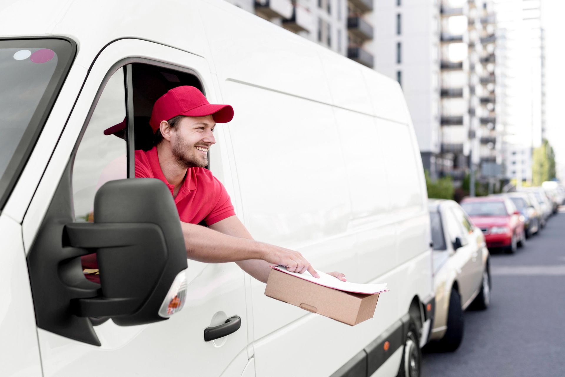 A smiling delivery person in a red uniform hands a package out of the driver's side window of a white van on a city street.