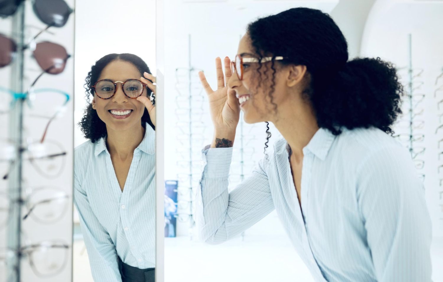 A person in a light blue shirt tries on glasses, smiling as they look at their reflection in an optical store mirror.
