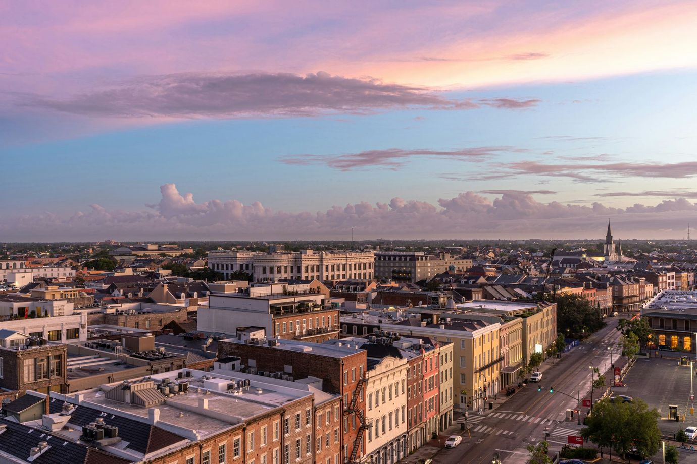An aerial view of the New Orleans French Quarter at sunset, showing historic buildings and a street under pink clouds.