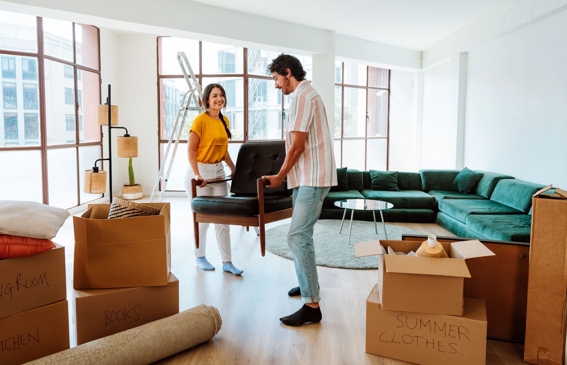 A couple carries an armchair into a bright, modern living room filled with cardboard moving boxes and a green sofa.