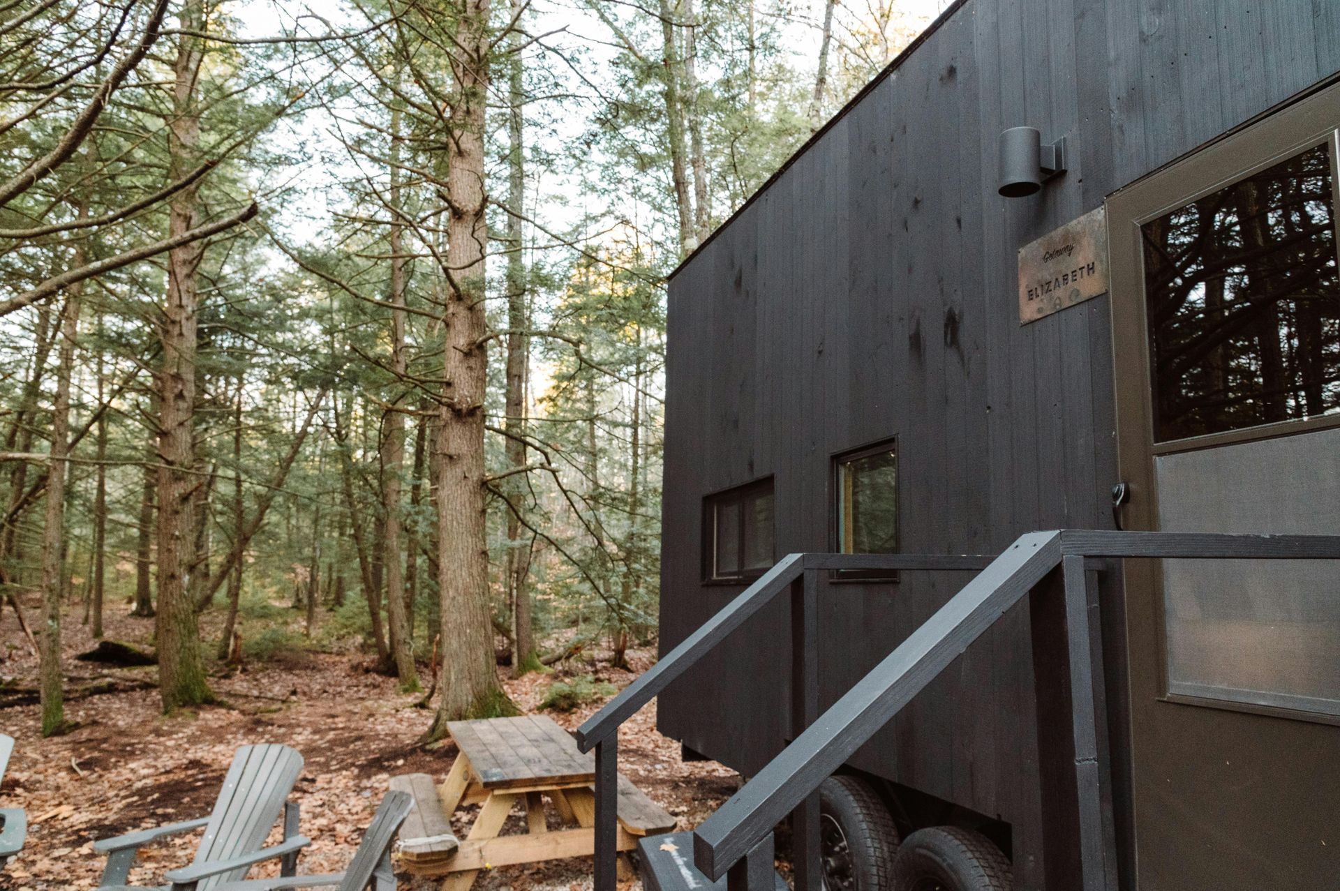 A small, modern black cabin sits in a dense forest, featuring a side entrance with stairs, a picnic table, and chairs.