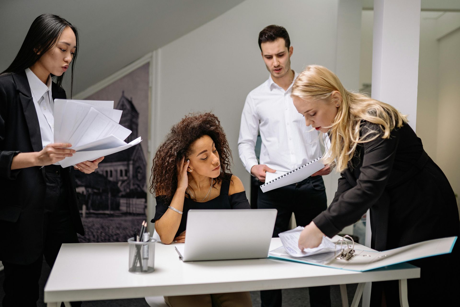 Four professionals collaborate around a white desk in an office, reviewing documents and working on a laptop.