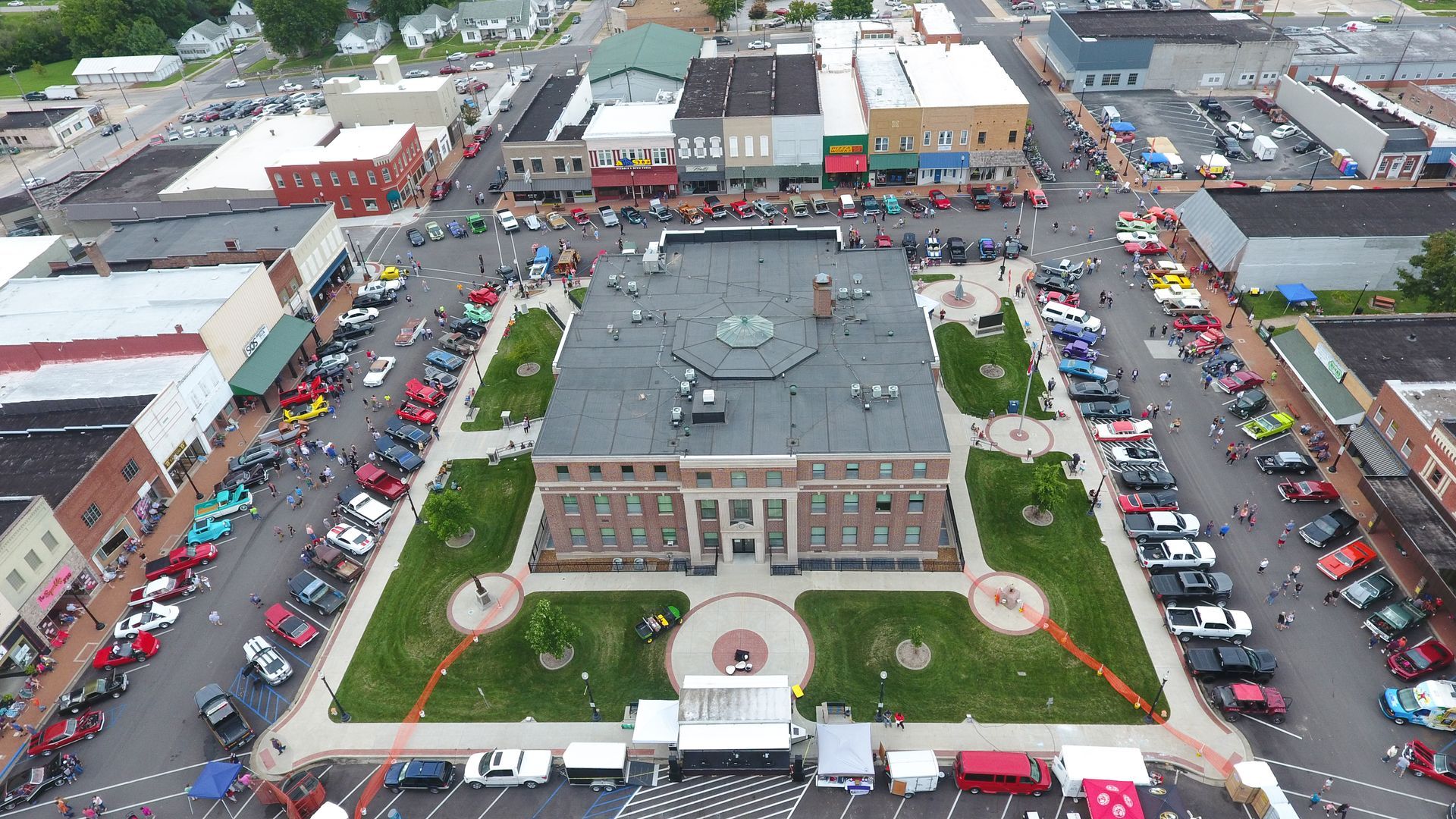 Aerial view of a town square with a brick building at its center surrounded by cars; event in progress.