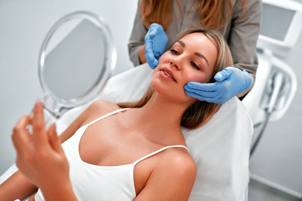 Woman having facial skin evaluation in a medical setting, holding a mirror and being touched by a gloved hand.