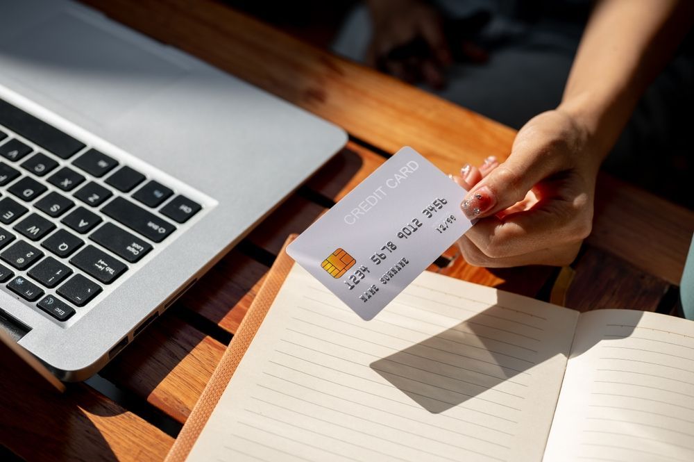 Person holding a credit card next to a laptop and a notebook on a wooden surface.