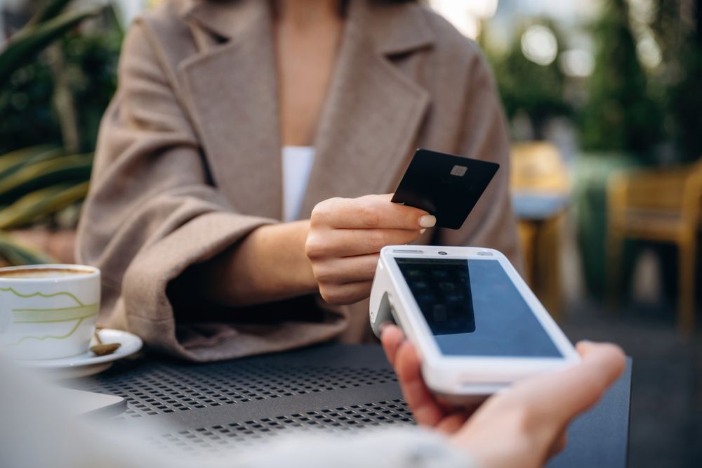 Person paying with a black card at a point-of-sale device on a patio.