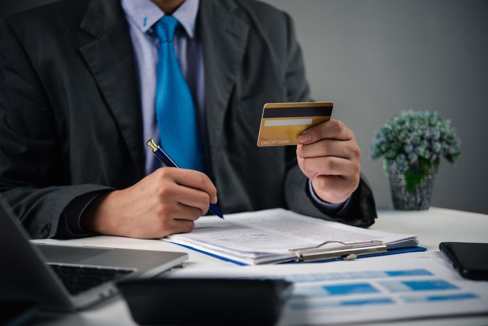 Man in suit holding credit card, writing on document at a desk.
