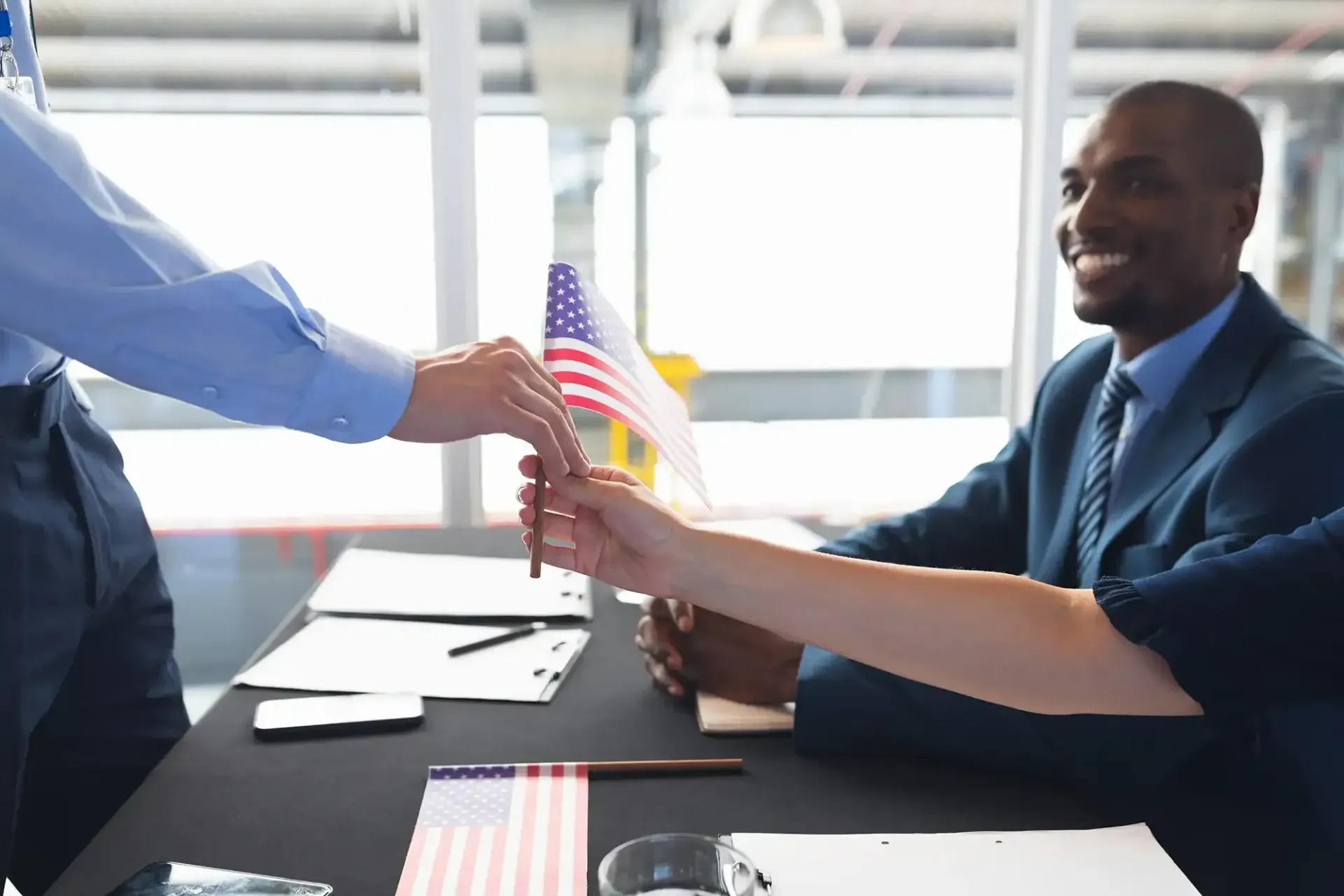 Two individuals exchange a pen across a desk featuring small American flags and documents in a professional setting.
