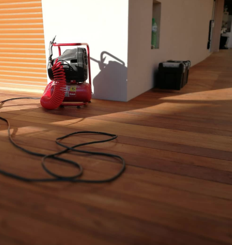 Un compressore d'aria rosso su un pavimento in legno accanto a una parete bianca, con cavi neri sparsi sul pavimento.