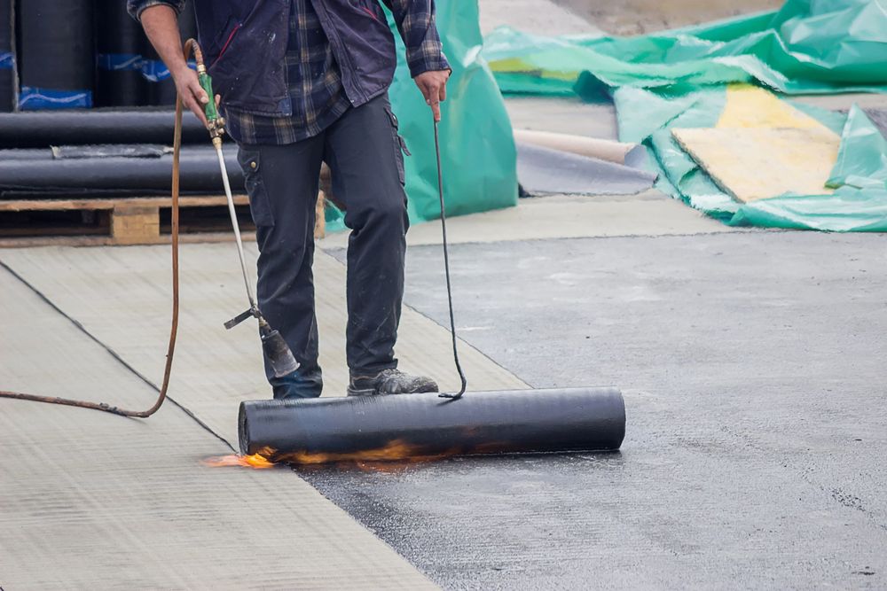 Un operaio utilizza una torcia per posare del materiale di copertura nero, con fiamme visibili sul tetto.