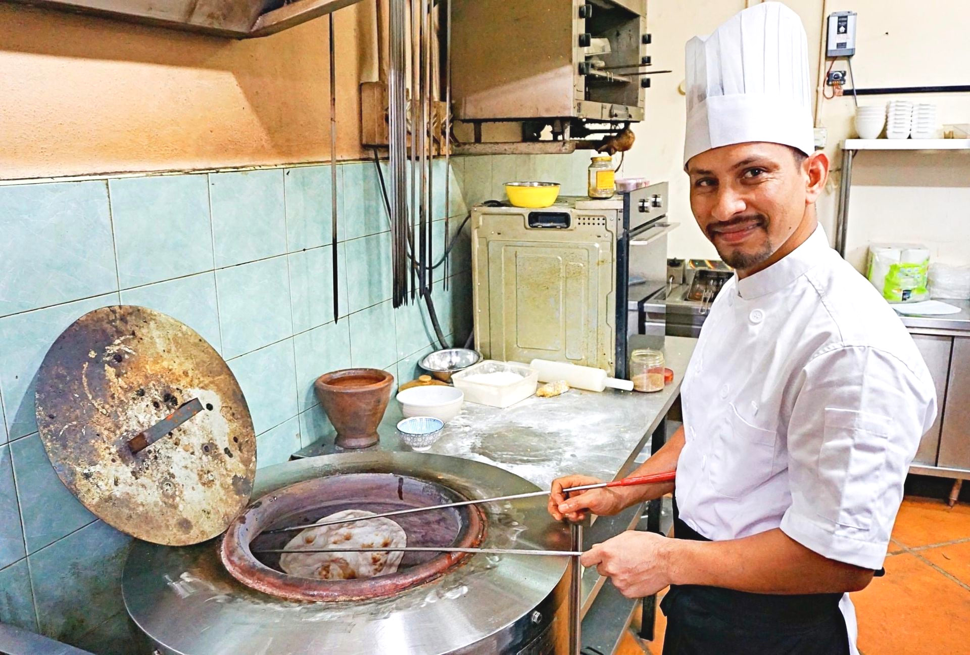 Chef stirring food in a tandoor clay oven beside a large tawa and stove