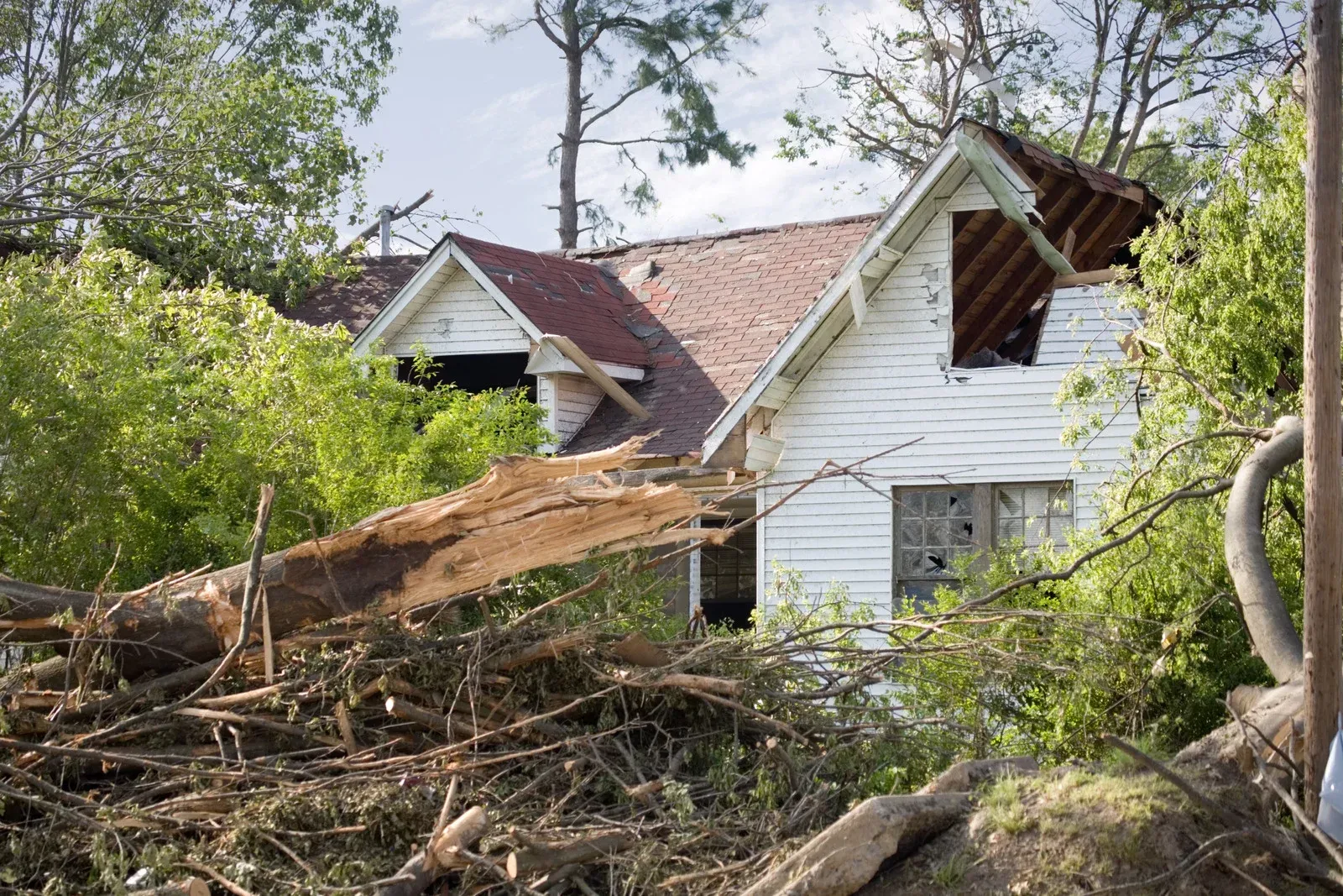 A white house with a damaged roof surrounded by debris and fallen trees after a storm.