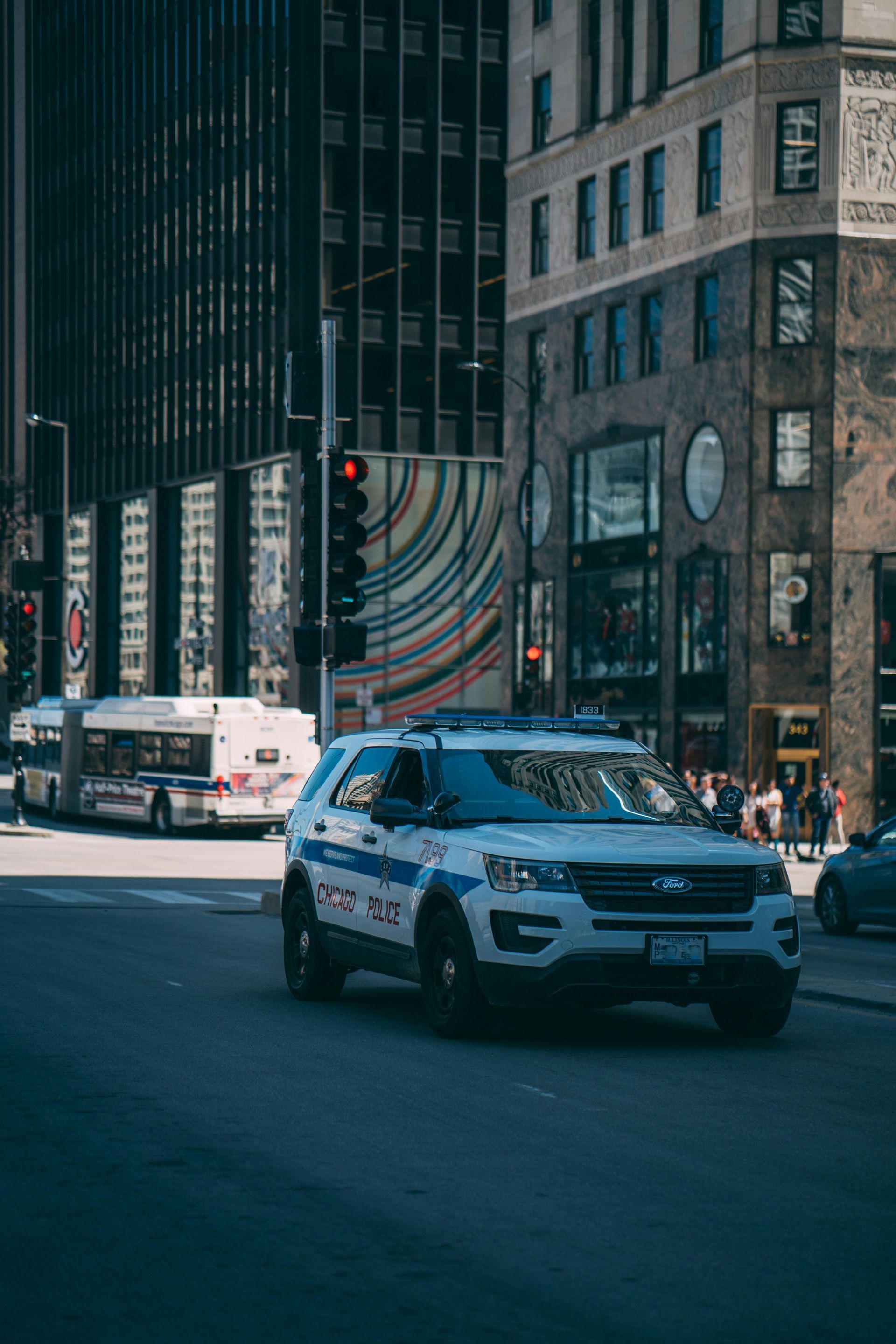 Police SUV driving on a city street, with a bus and buildings in the background under a sunny sky.