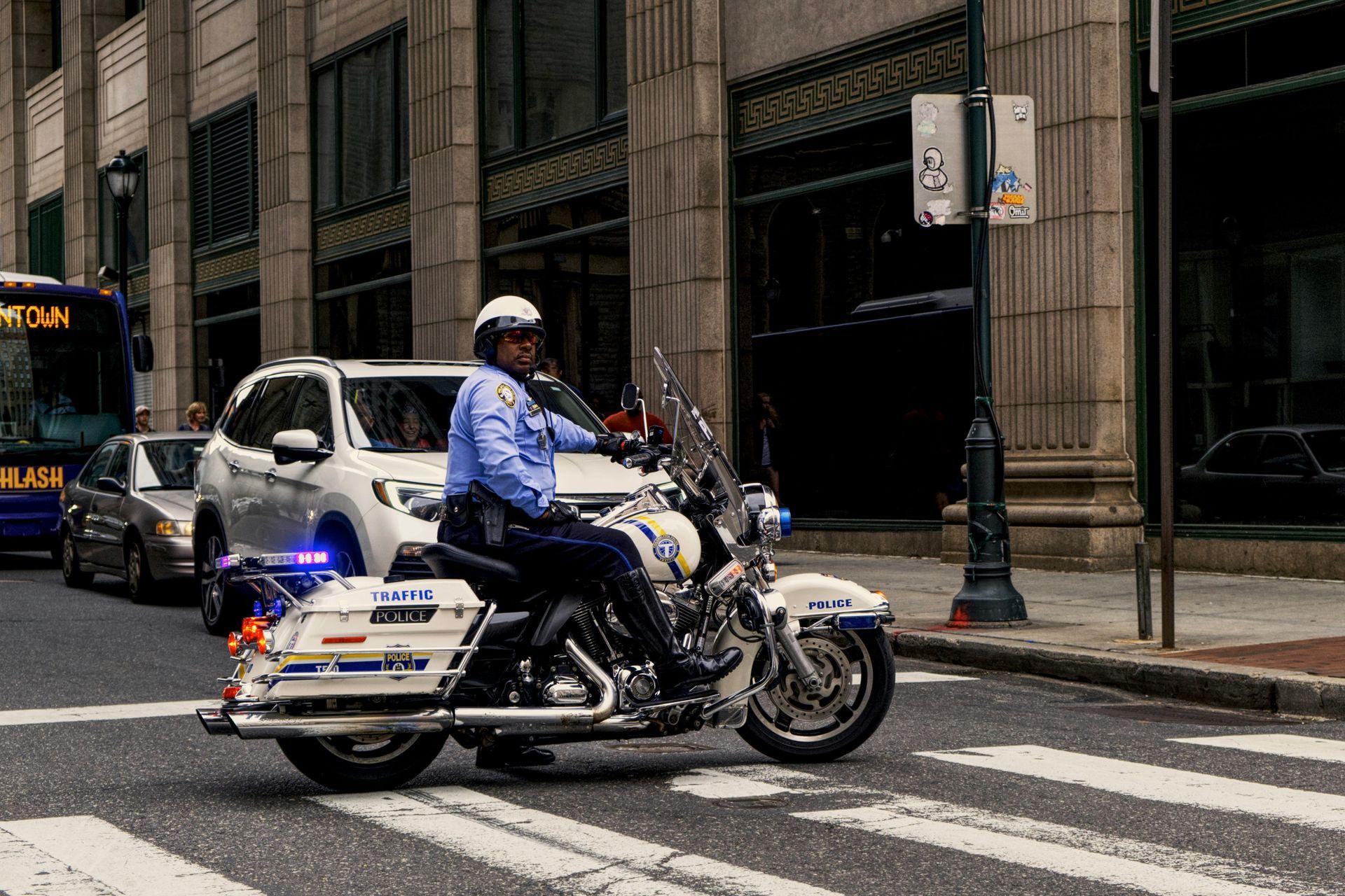 Police officer on motorcycle in a city street, with lights flashing. Buildings and traffic are in the background.