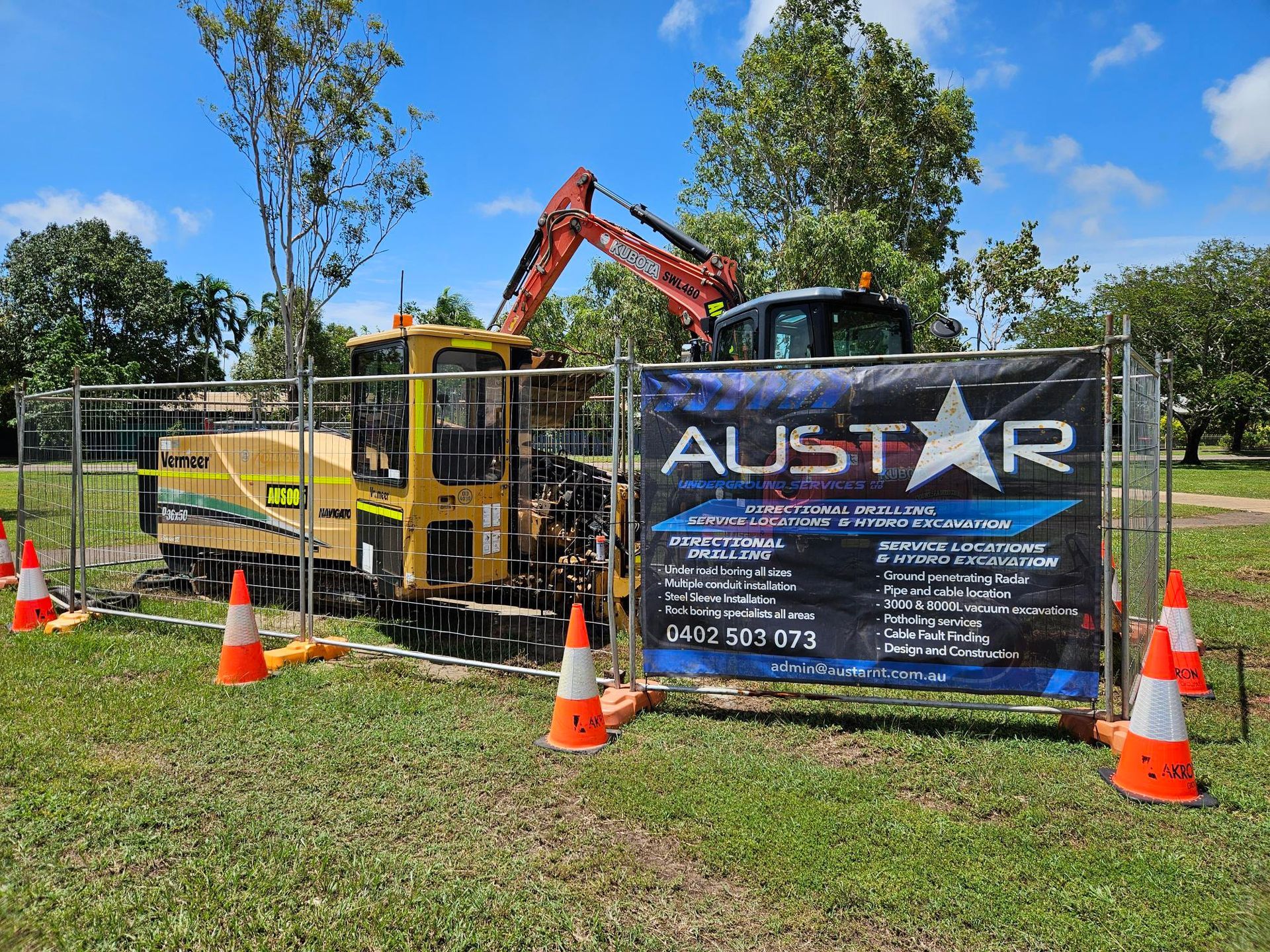 Construction site with a yellow drilling machine behind a fence; orange cones and a banner with