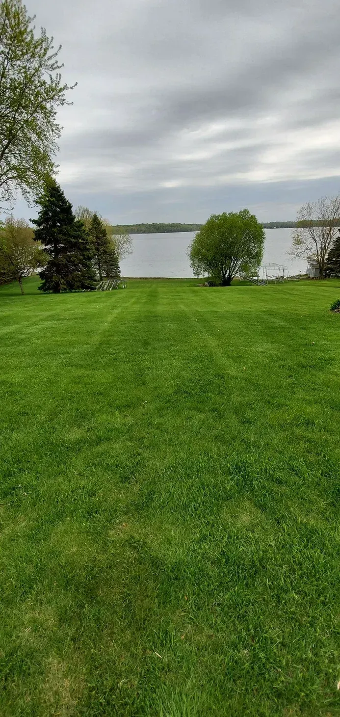 Green lawn leads to a lake under a cloudy sky, with trees on either side.