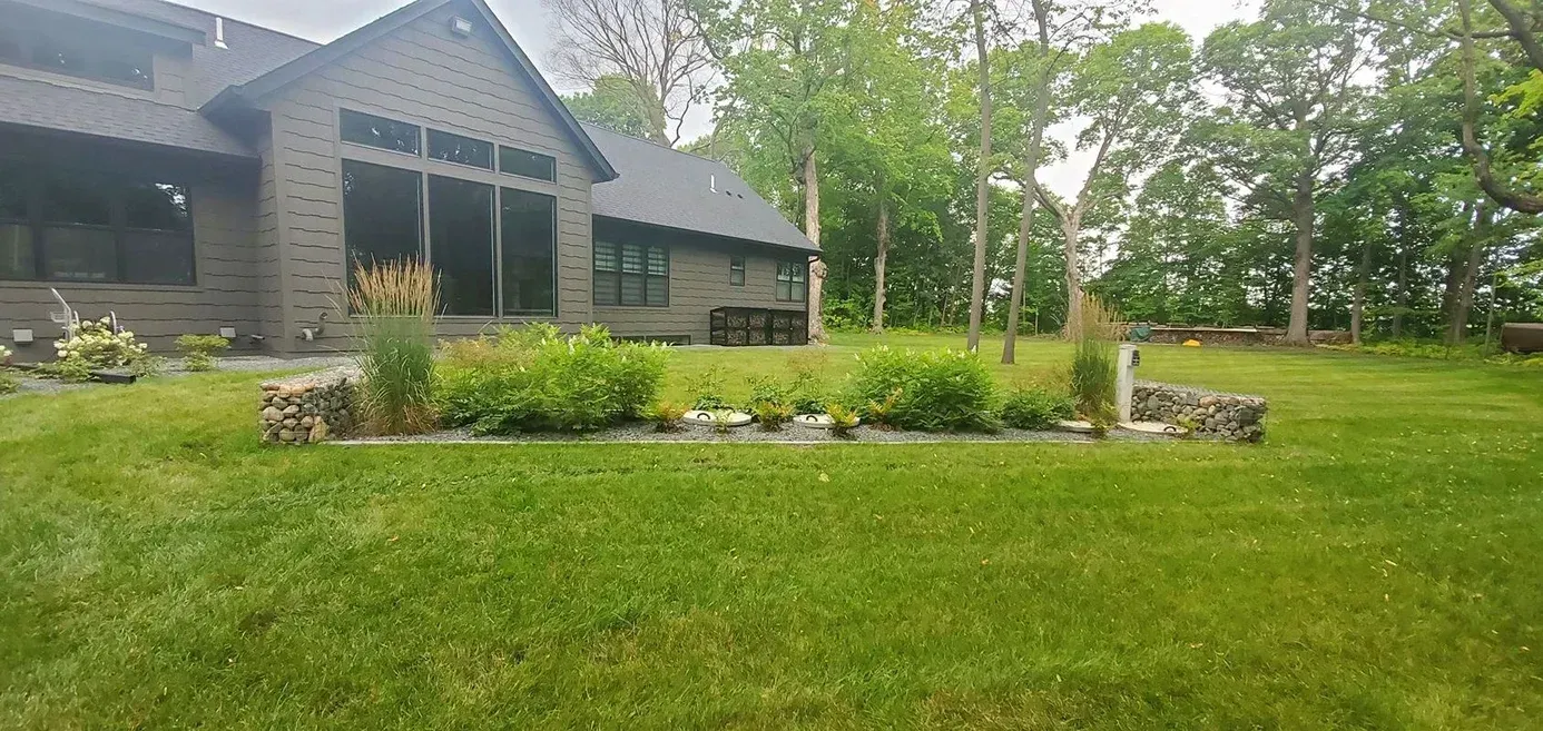 A house with a dark roof and large windows overlooks a green lawn and garden. Trees frame the background.