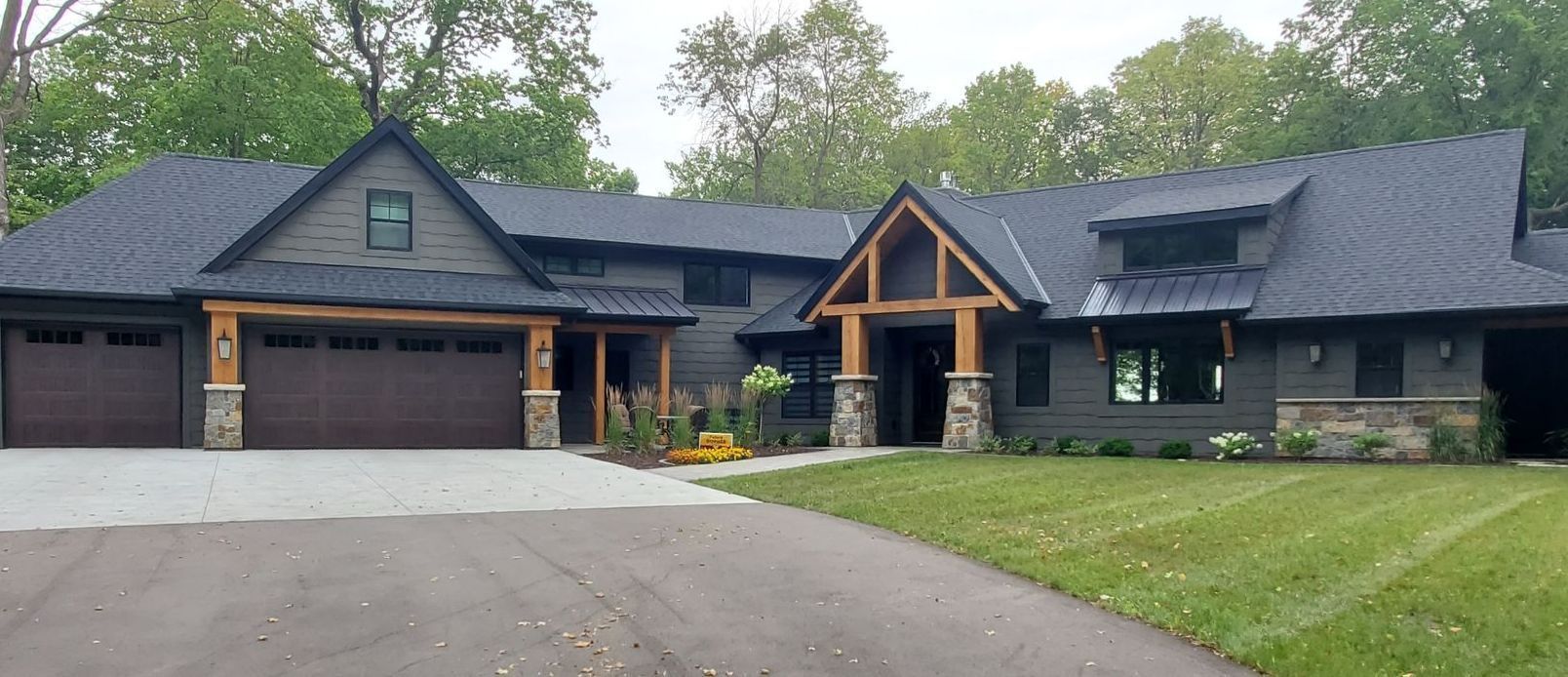 Gray house with brown garage doors and a stone and wood facade, set in a green yard with a paved driveway.