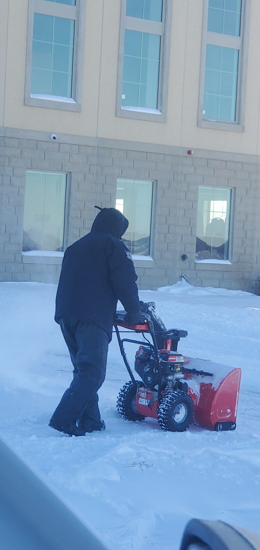 Person using a snowblower on a snow-covered area in front of a building with windows.
