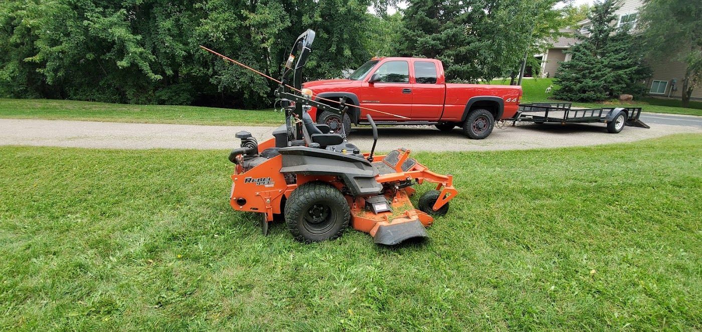 Orange lawn mower on green grass near a red truck and trailer.