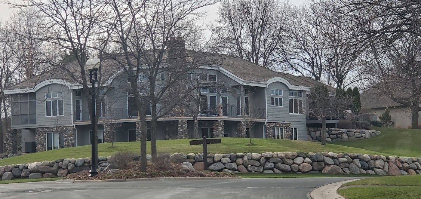 Large stone house on a hill, surrounded by trees and a rock wall. Overcast sky.